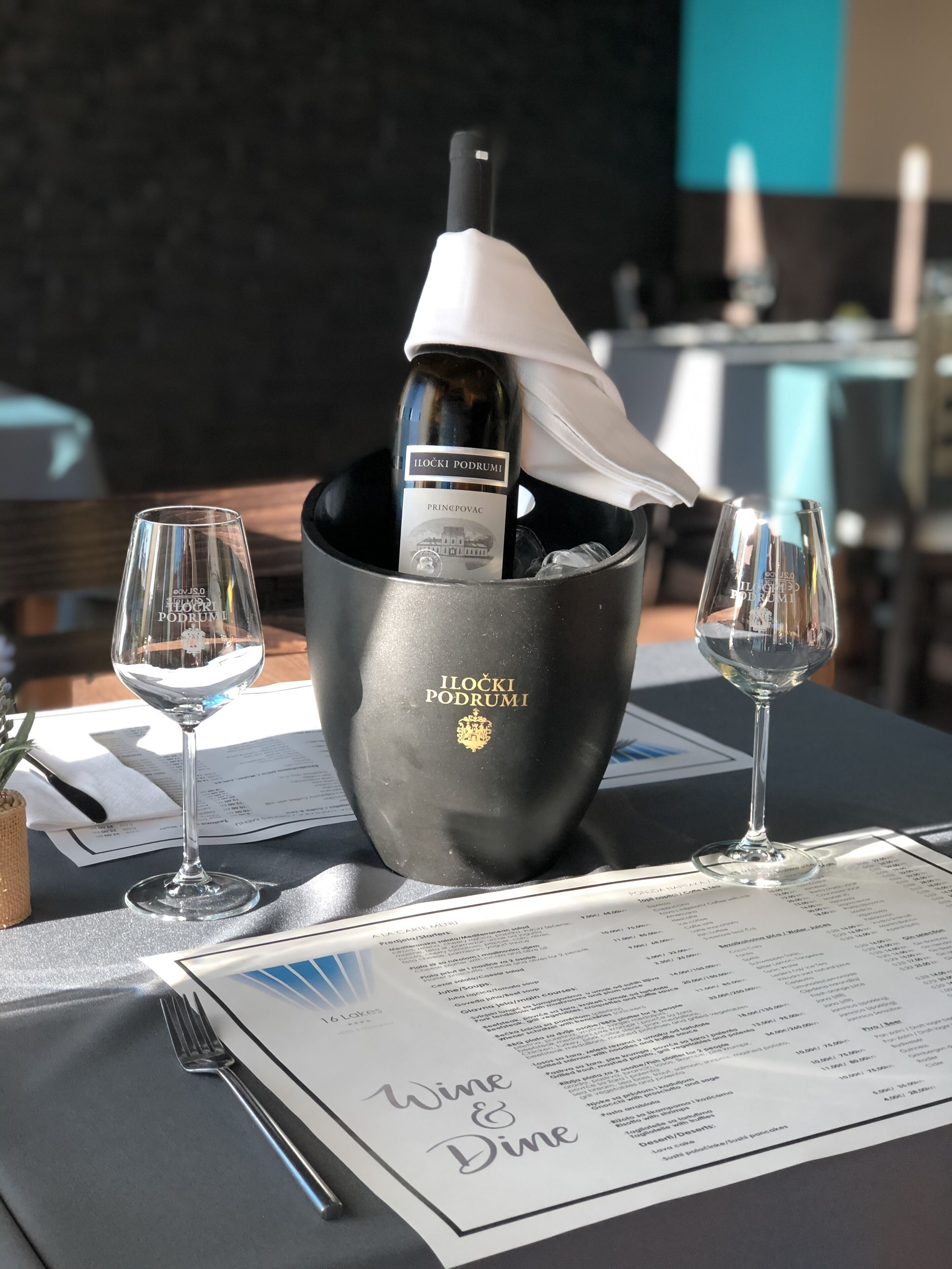 Une table avec un menu, deux verres à vin et une bouteille de vin Iłocki Podrumi dans un seau noir.