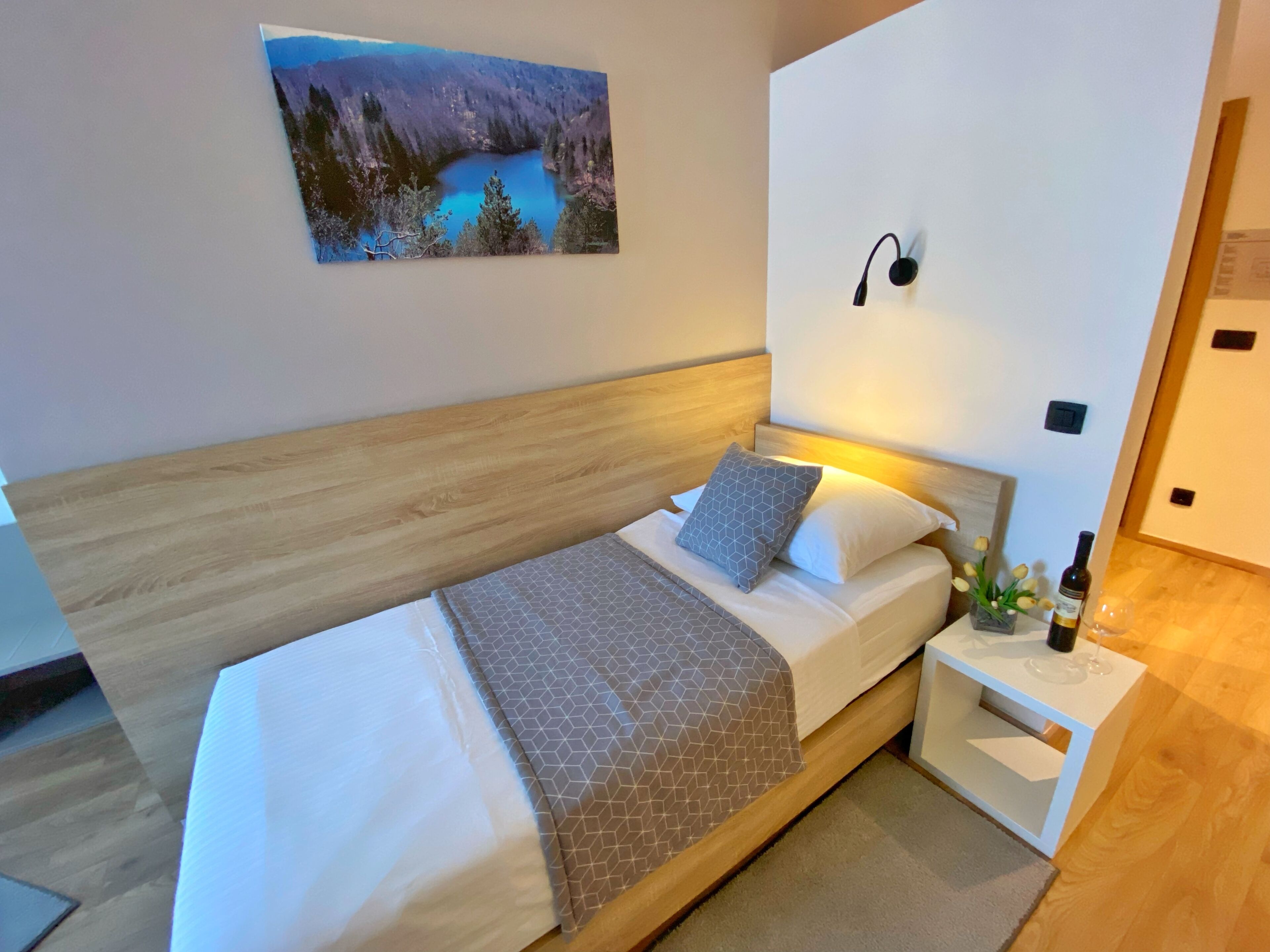 A modern bedroom with a wooden headboard, a bed with white bedding and patterned pillows, a nightstand with a bottle and glass, and a painting of a lake on the wall.