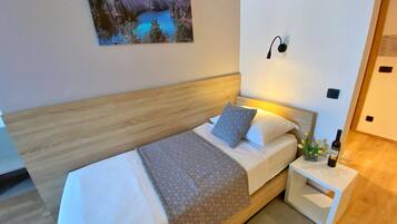 A modern bedroom with a wooden headboard, a bed with white bedding and patterned pillows, a nightstand with a bottle and glass, and a painting of a lake on the wall.