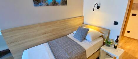 Un dormitorio moderno con cabecera de madera, cama con ropa de cama blanca y almohadas estampadas, una mesita de noche con una botella y un vaso, y un cuadro de un lago en la pared.