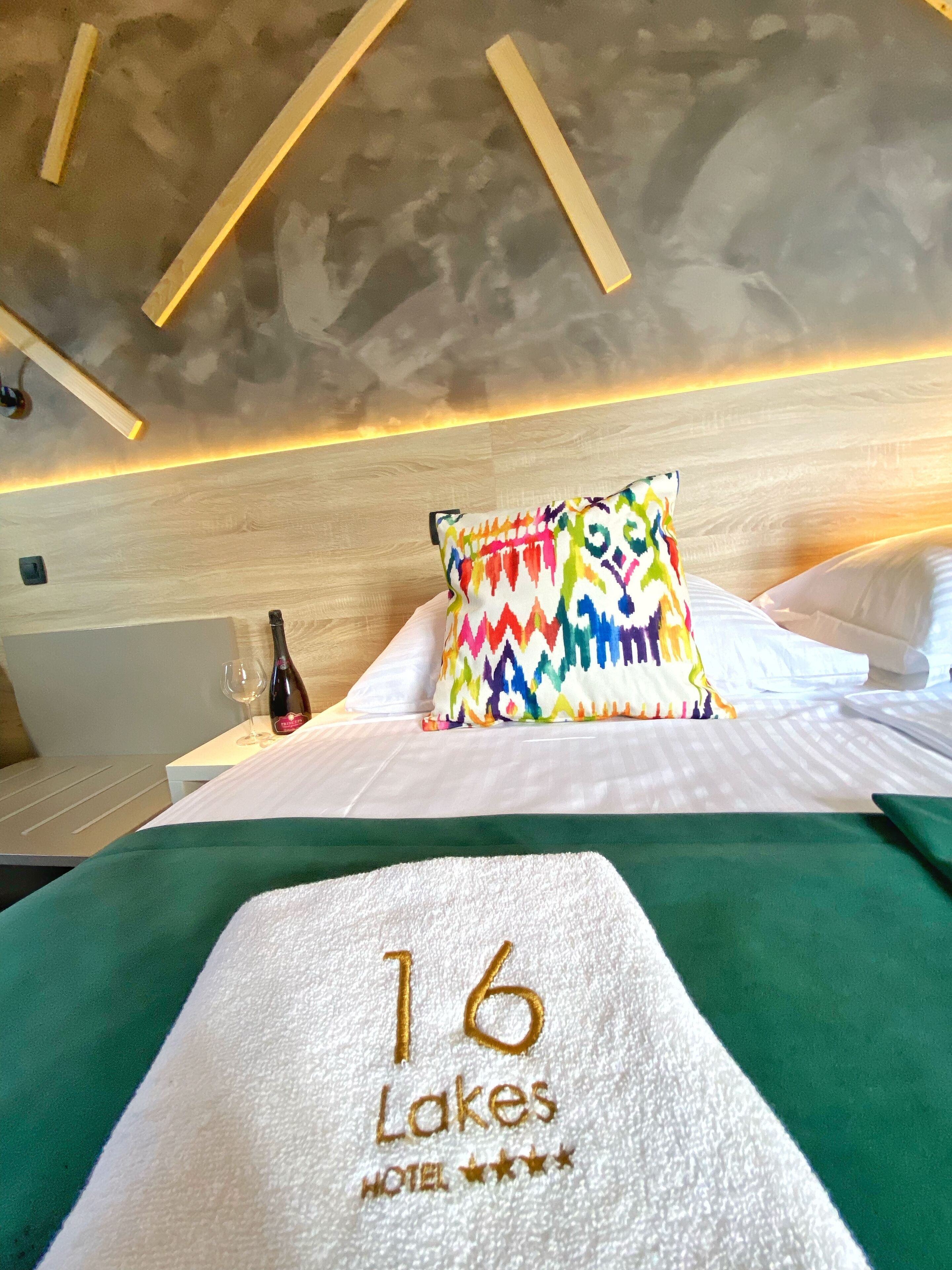 Ein Hotelzimmer mit Bett, farbigem Kopfkissen, einer Weinflasche und einem Handtuch mit dem Schriftzug '16 Lakes Hotel'.