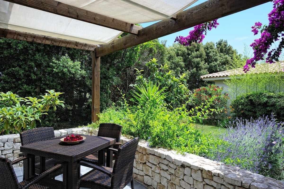 An outdoor patio with a table and chairs, surrounded by a stone wall and lush greenery.
