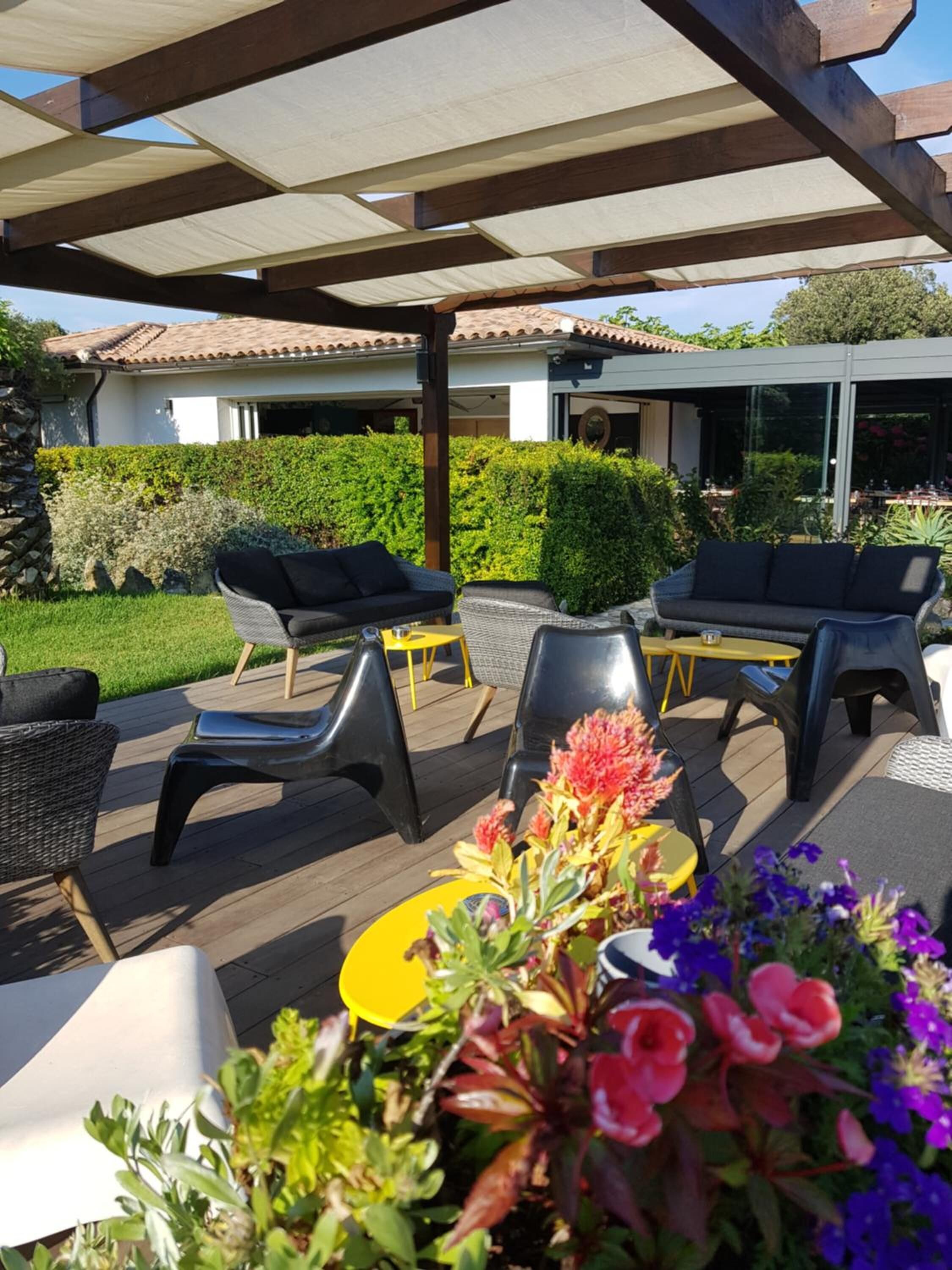 Un patio al aire libre con mobiliario de mimbre y una mesa amarilla, rodeado de follaje y un edificio al fondo.