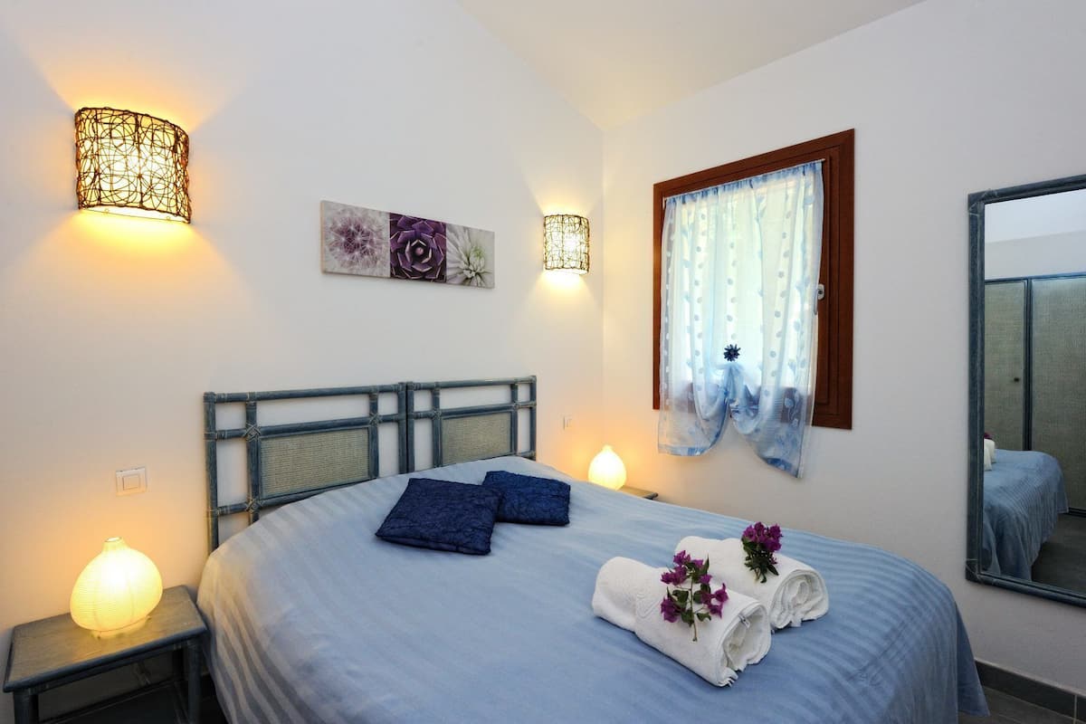 A neatly made bed with blue bedding, two wall sconces, a framed artwork, a window with sheer curtains, and a mirror on the wall.