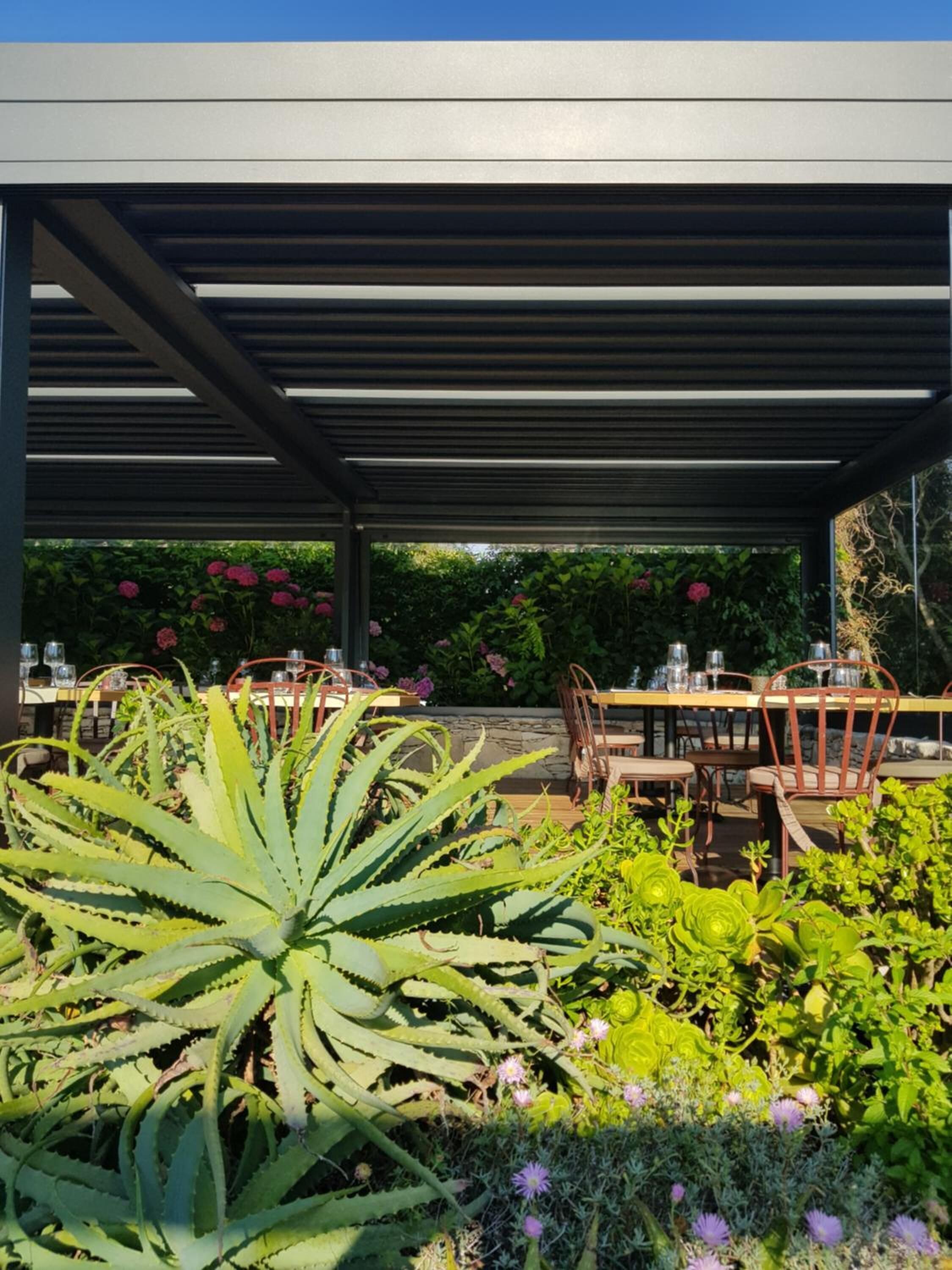 An outdoor patio with tables and chairs, surrounded by various plants and flowers.