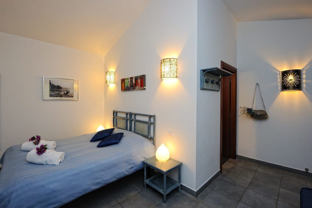 A bedroom with a bed, bedside lamps, a small table, a door, and wall-mounted lights.