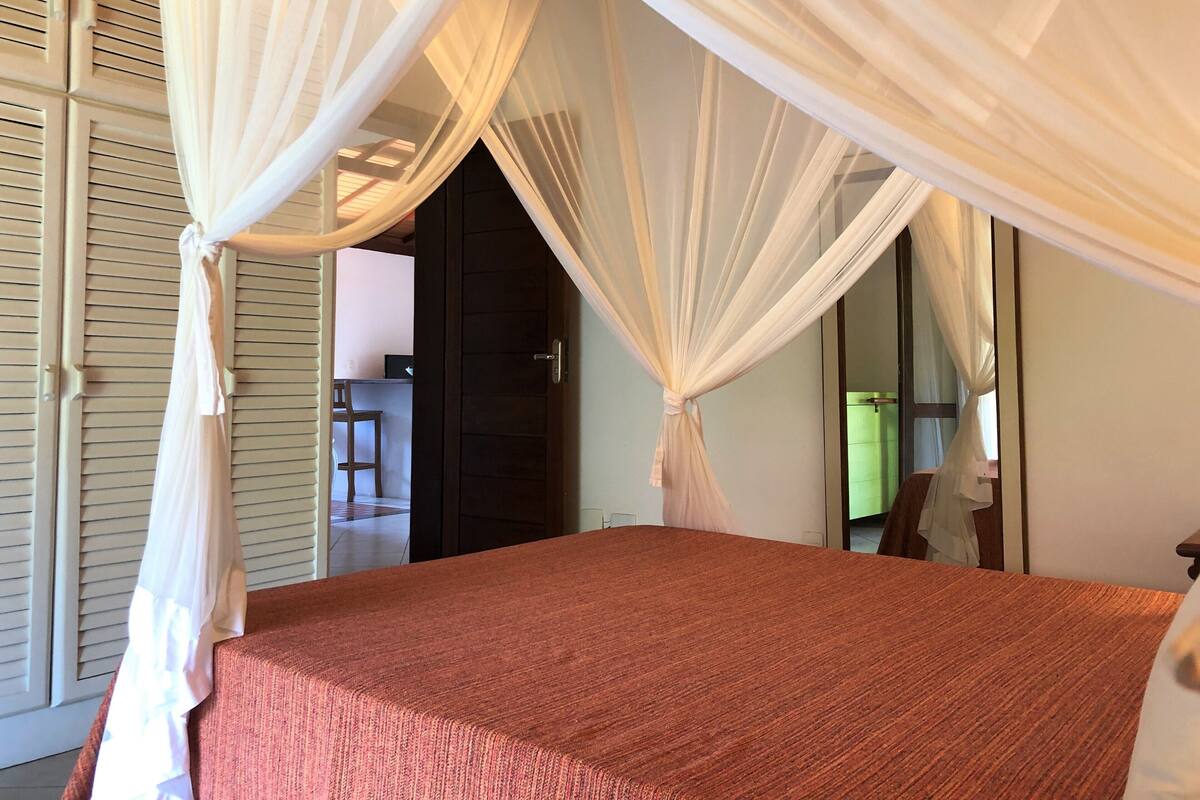 A bedroom with a bed, a window with sheer curtains, and a door leading to another room.