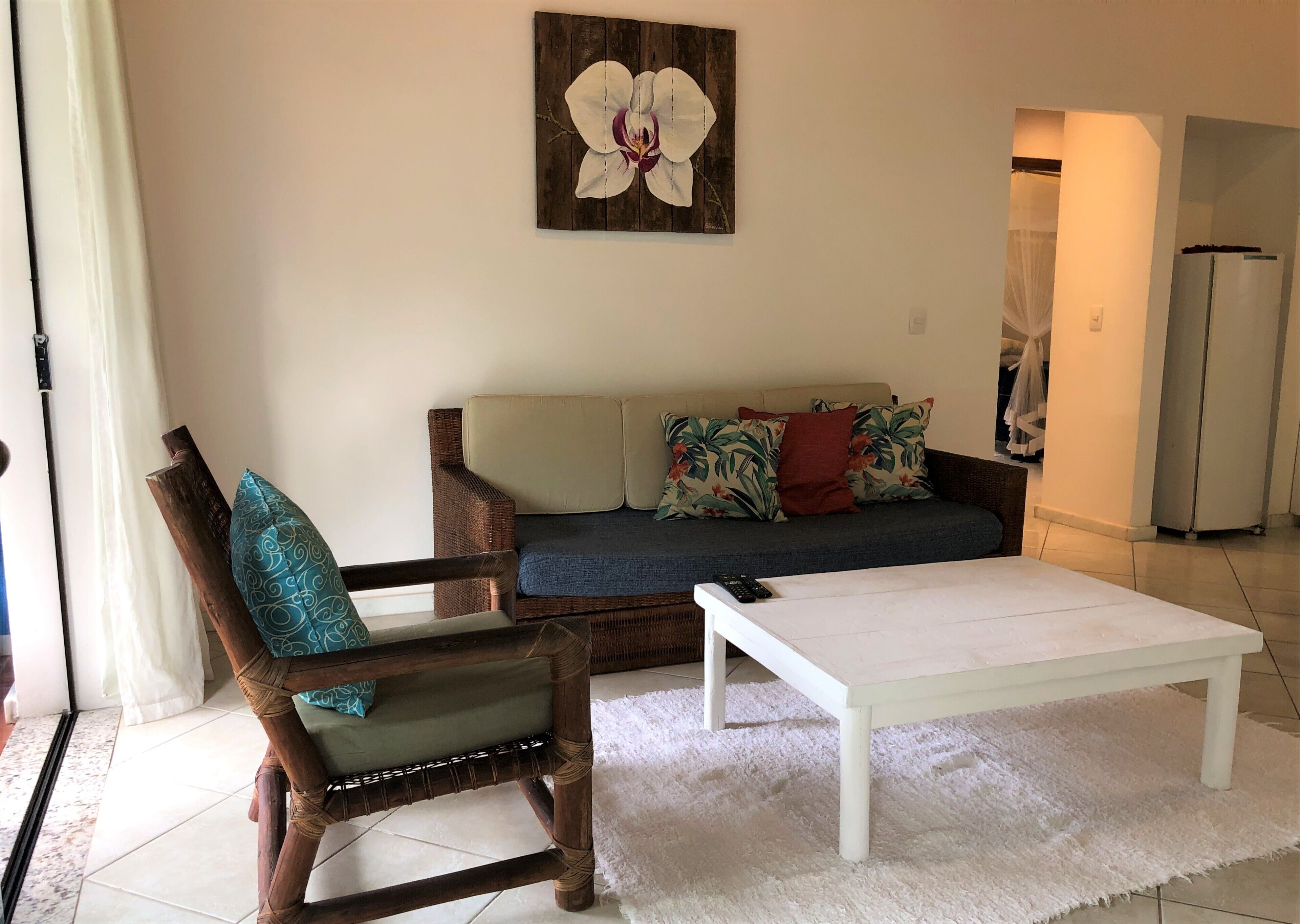 Una sala de estar con un sofá, una silla de madera con cojín, una mesa de centro blanca y una pintura de una orquídea en la pared.