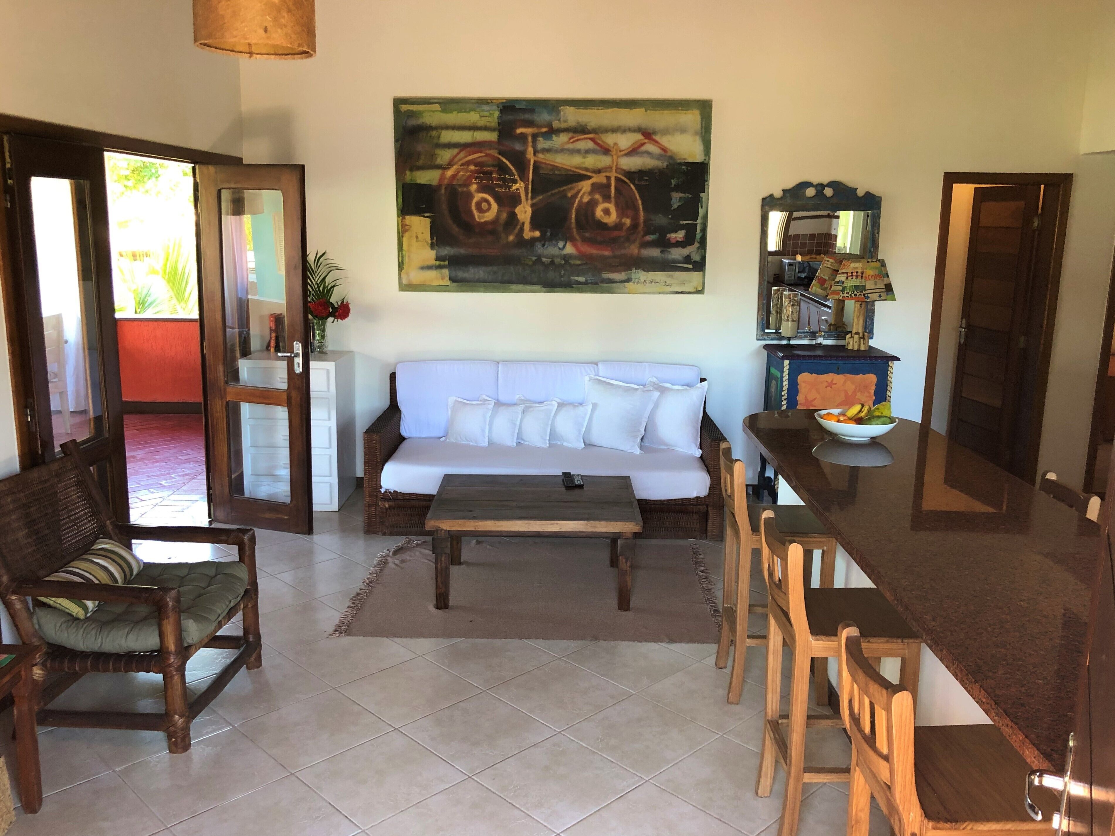 Ein Wohnzimmer mit einer Couch, einem Couchtisch, Holzstühlen, einem Esstisch mit Stühlen, einem Gemälde an der Wand und einer Küchenzeile mit Kühlschrank und Mikrowelle.