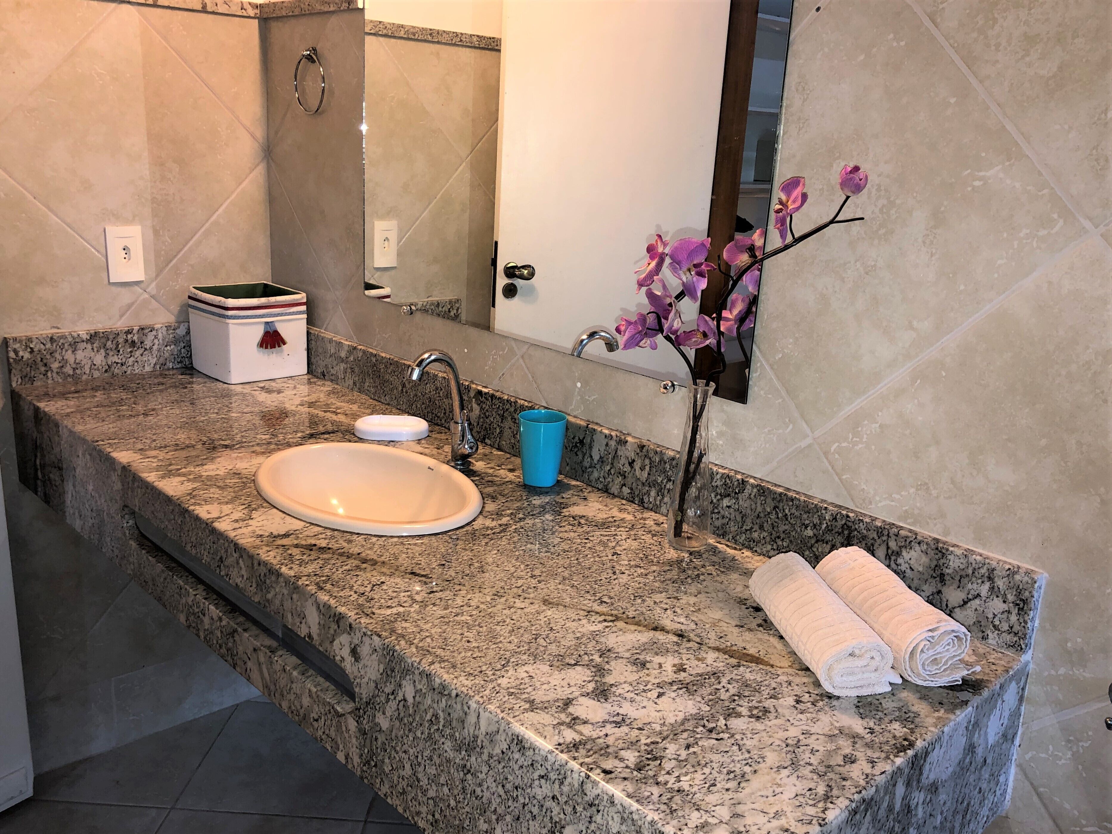 Une salle de bain avec un comptoir en granit, un lavabo blanc, une tasse bleue et un vase contenant des orchidées violettes.