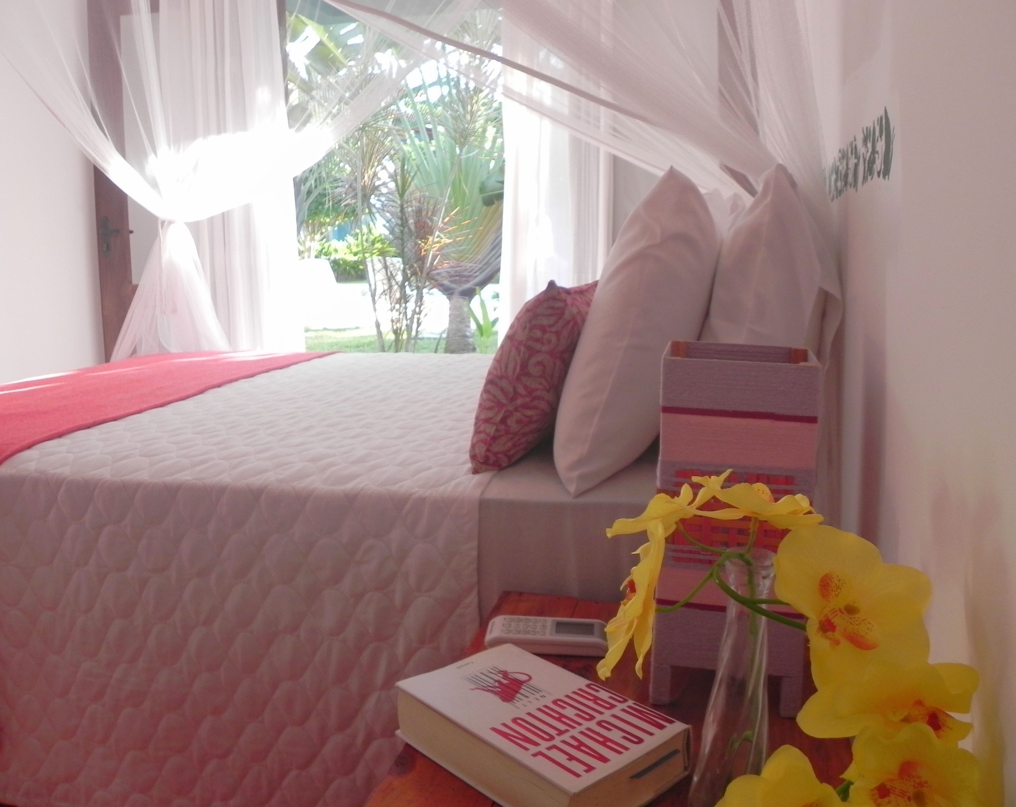 Quarto com cama, mesa de cabeceira com um livro, vaso com flores e janela com cortinas.