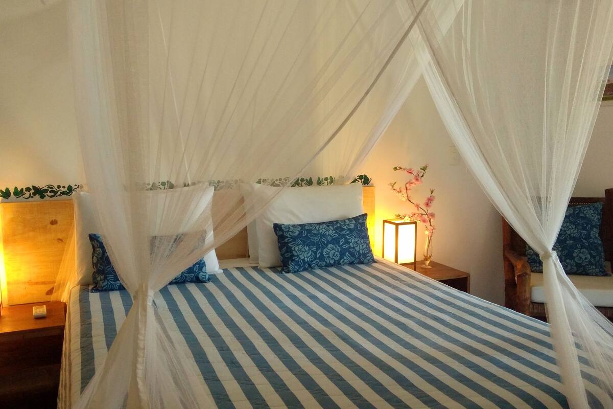 A bed with a striped bedspread and blue pillows, a wooden nightstand with a lamp, and a chair with a floral pillow.