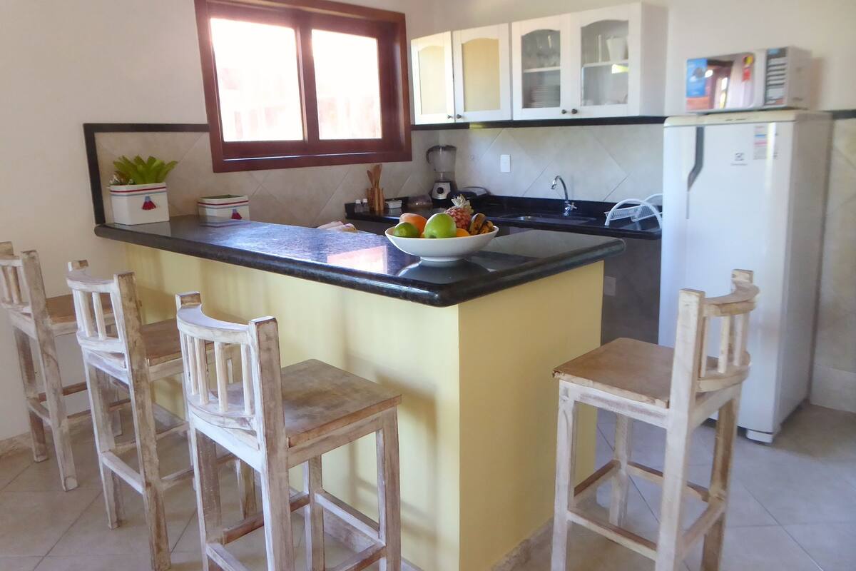 A kitchen with a central island, wooden chairs, a bowl of fruit, and a refrigerator.