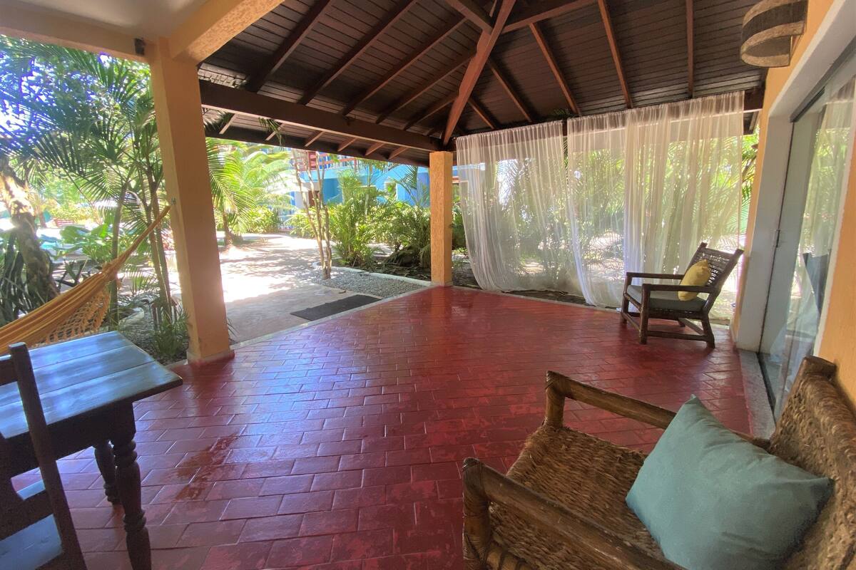 A covered outdoor area with a red brick floor, wooden furniture, and a view of a garden.