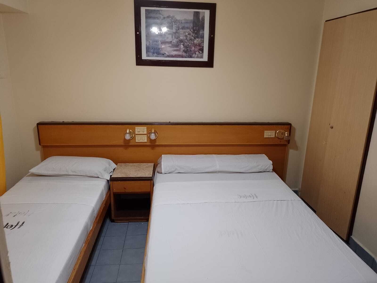 A hotel room with two single beds, a wooden headboard, a small bedside table with a lamp, and a framed picture on the wall.
