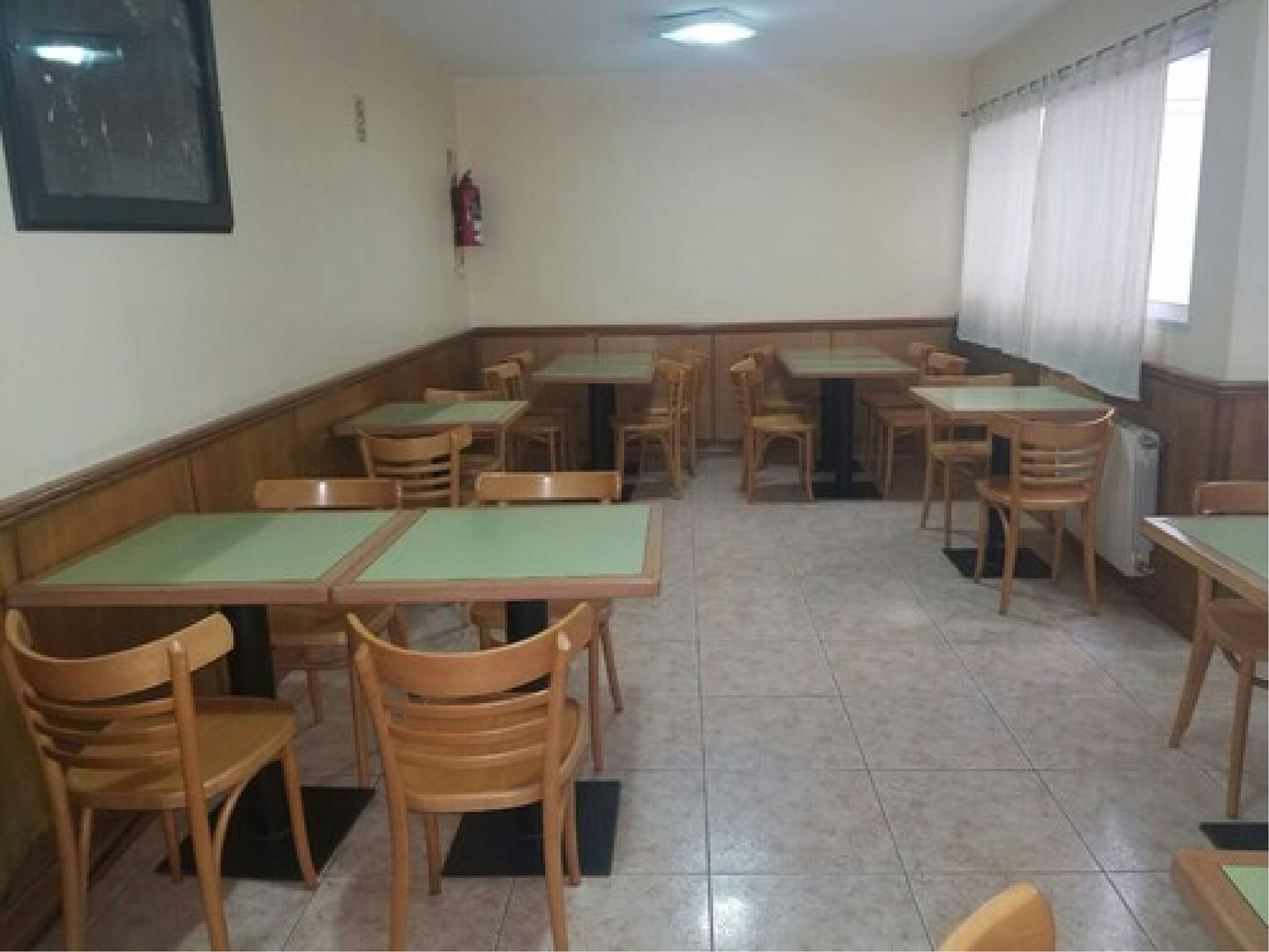 Un comedor con varias mesas y sillas, un extintor en la pared y una ventana con cortinas.