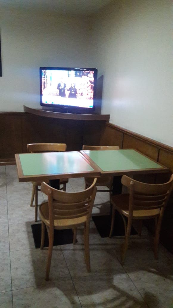 Una habitación con una televisión sobre un mueble bajo, dos mesas de madera con sillas, y un piso de baldosas.