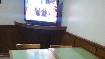Una habitación con una televisión sobre un mueble bajo, dos mesas de madera con sillas, y un piso de baldosas.