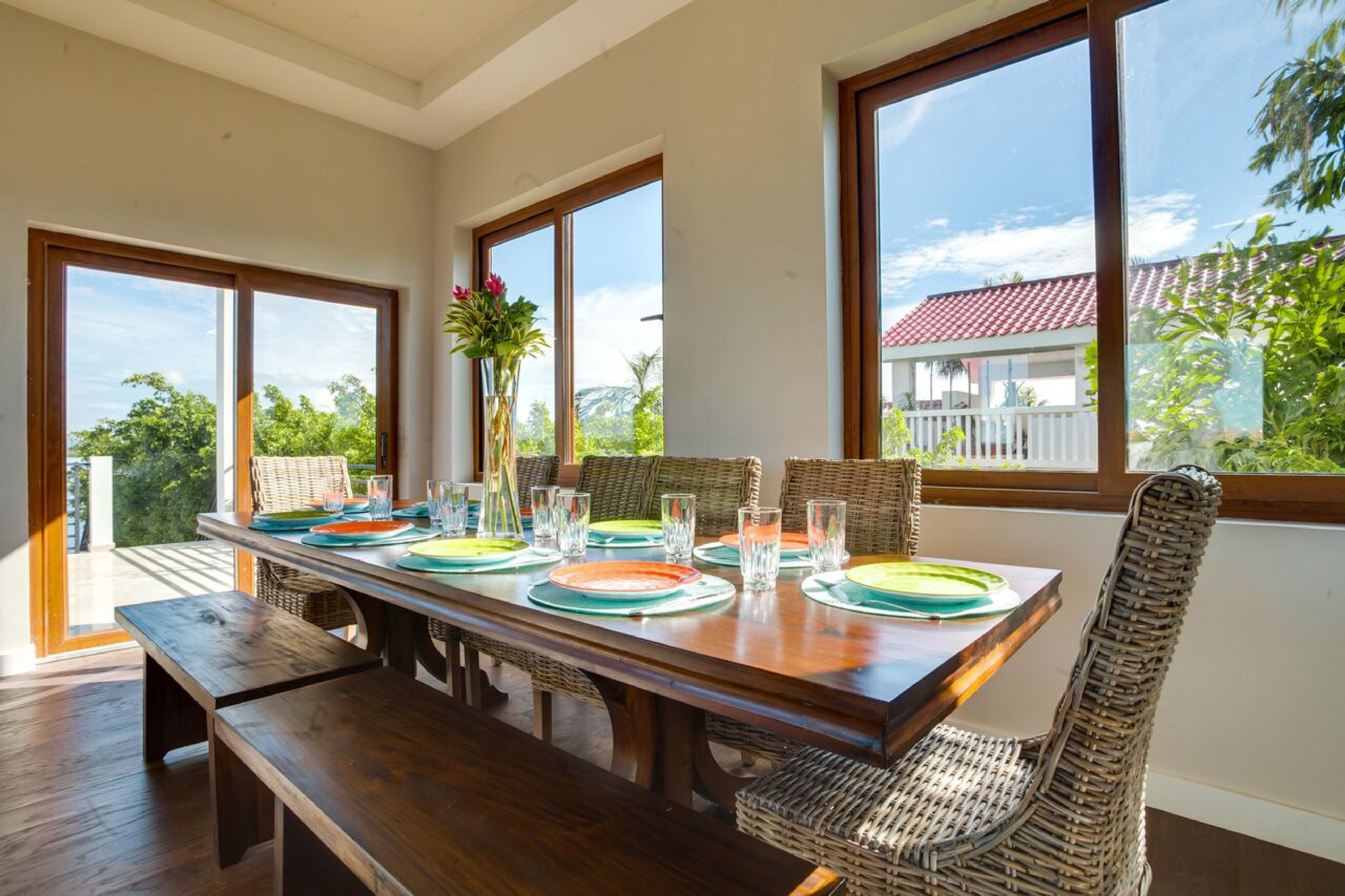 Un coin repas avec une table en bois dressée pour quatre personnes, entourée de chaises en osier, de grandes fenêtres donnant sur un paysage verdoyant et un bâtiment avec un toit rouge.