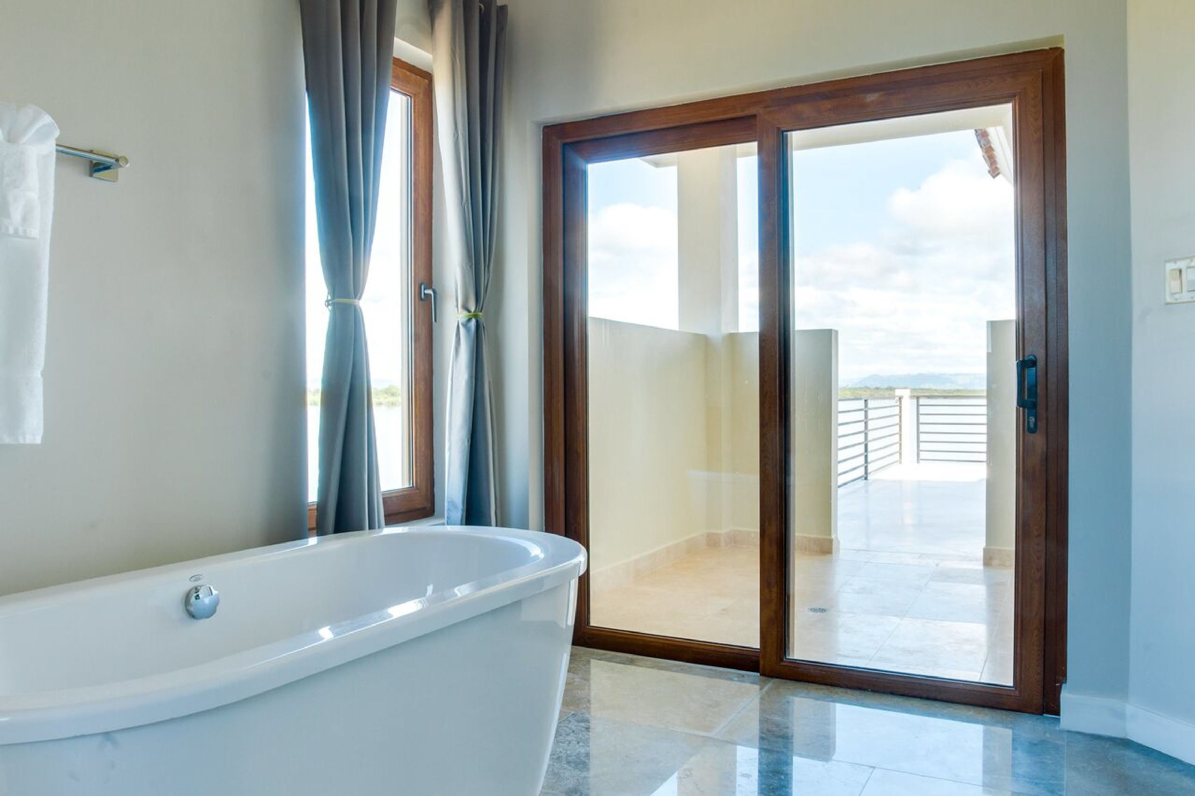 A bathroom with a bathtub, a window with curtains, and a sliding glass door leading to an outdoor balcony.