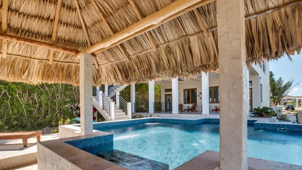 A pool area with a thatched roof, a staircase leading to a covered deck, and a view of a modern building in the background. - Villa Laguna Gecko (Placencia)