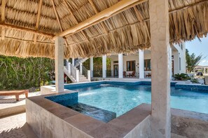 A pool area with a thatched roof, a staircase leading to a covered deck, and a view of a modern building in the background.