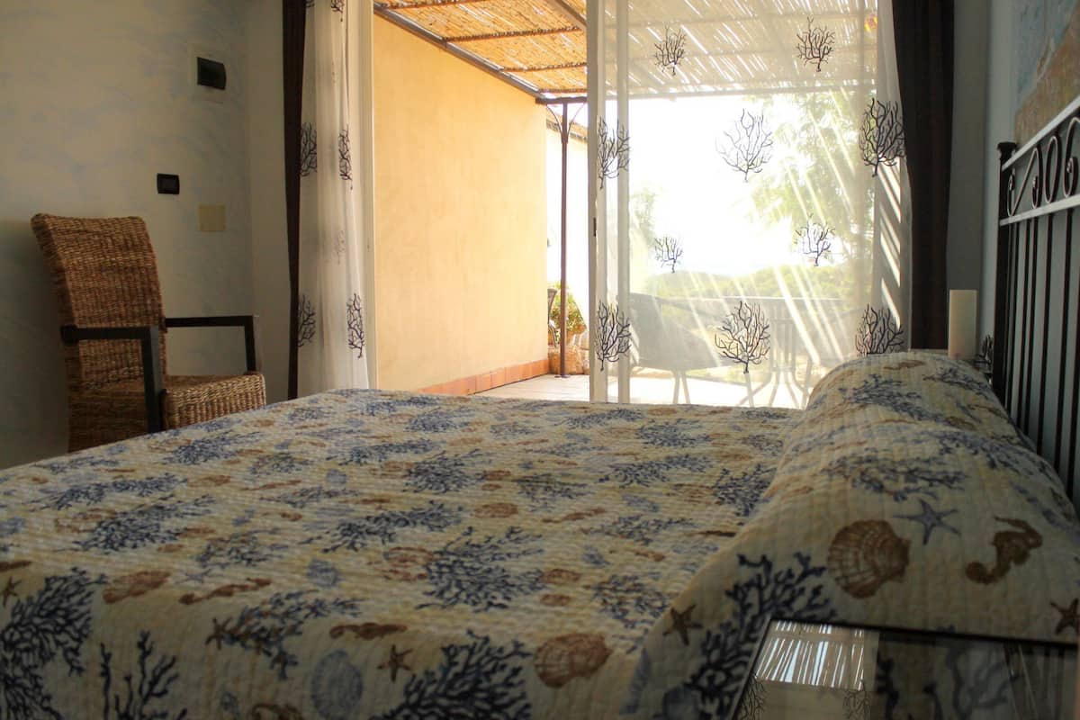 A bedroom with a bed, a wicker chair, and a view of the outdoors through a glass door.