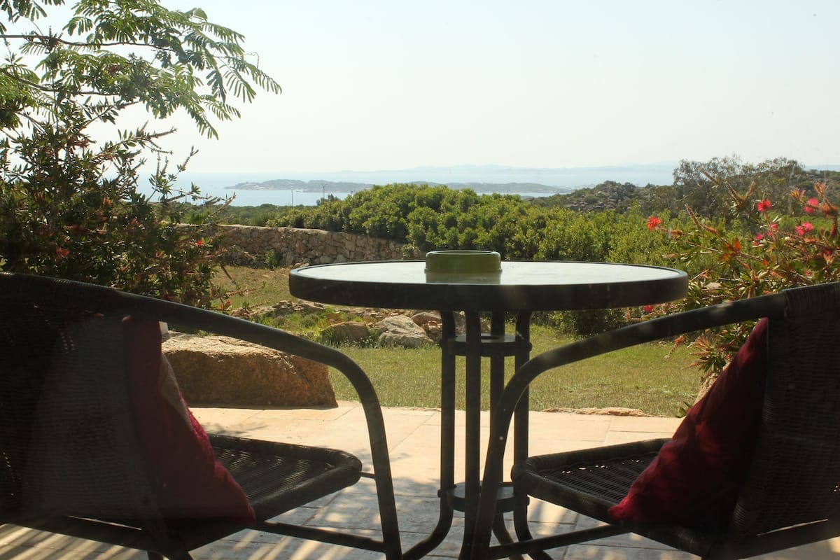 An outdoor seating area with a round table and two chairs, overlooking a scenic landscape with greenery and distant hills.