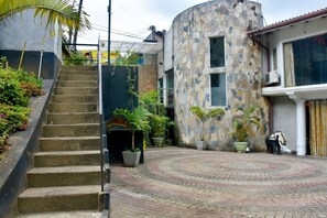 Um edifício curvo com fachada de pedra, uma escadaria que leva a uma entrada com portão e um pátio pavimentado com plantas em vasos.