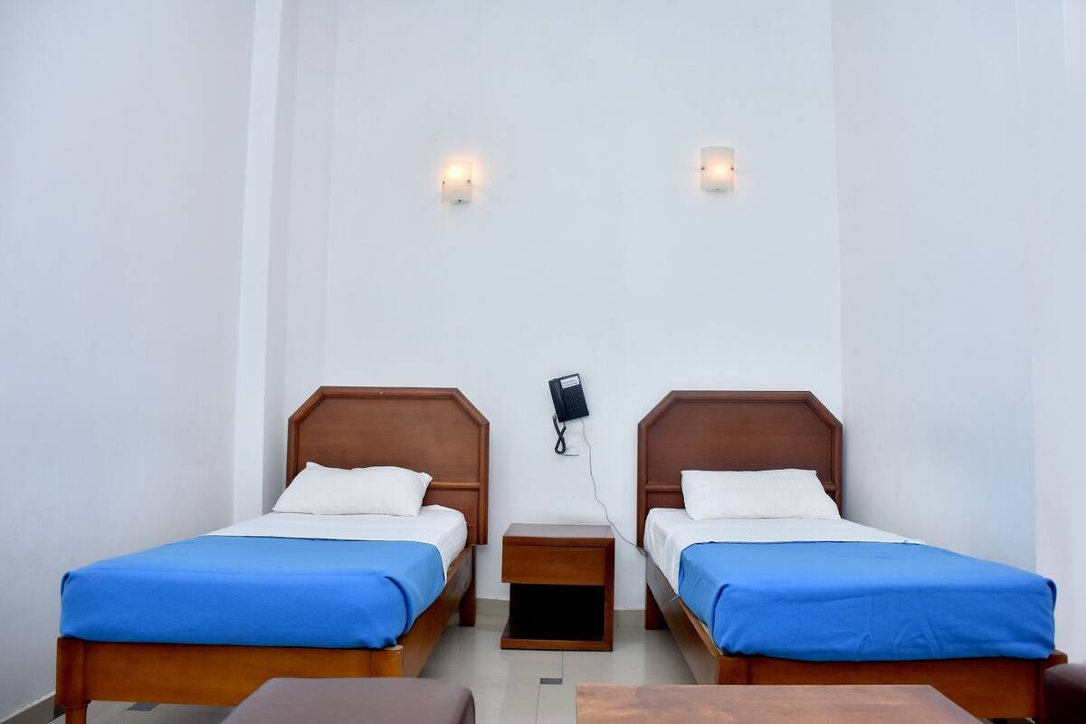 A room with two wooden beds, each with a blue bedspread, a small wooden nightstand between them, and a wall-mounted telephone.