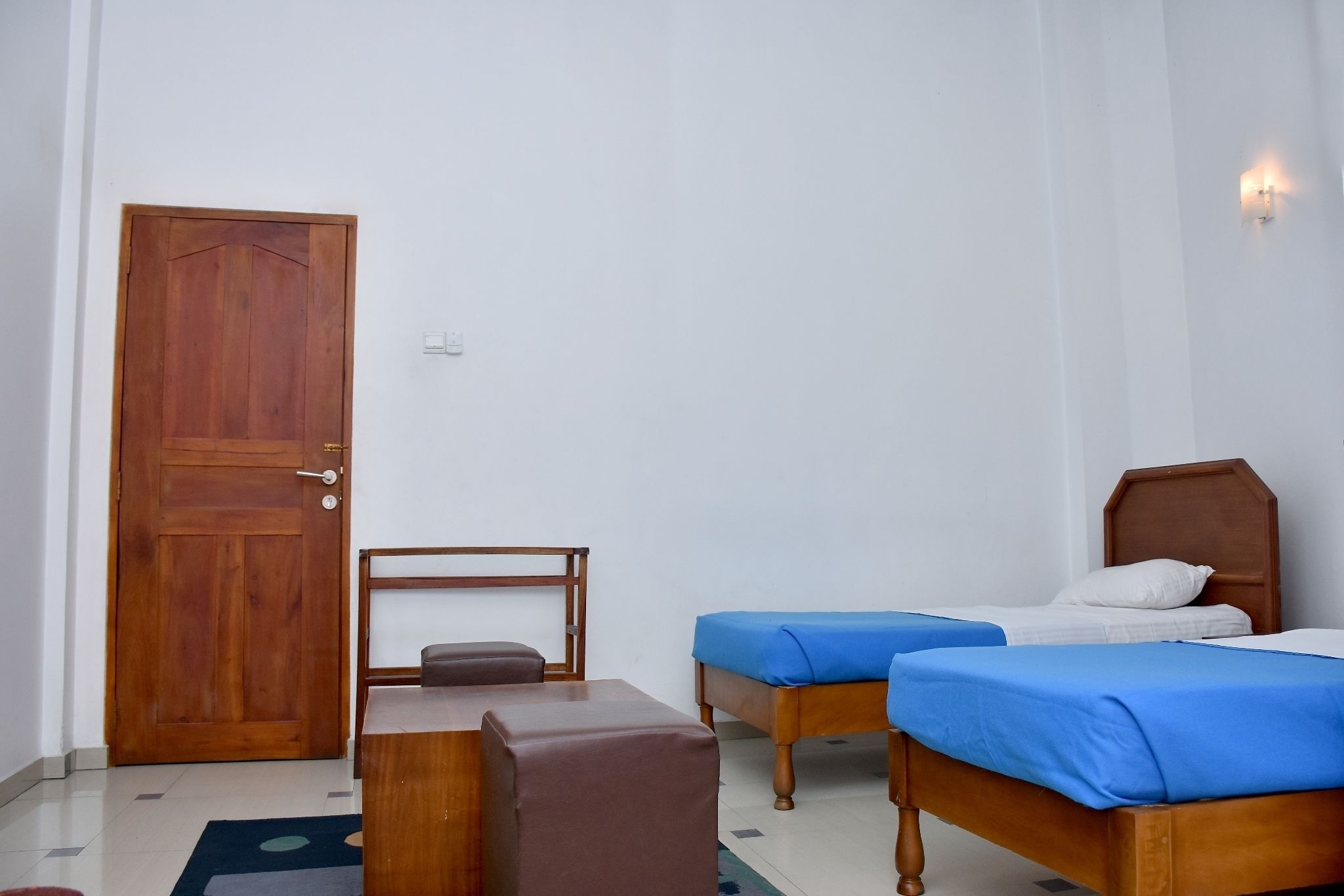 Una habitación con dos camas, una puerta de madera, una mesita pequeña y una lámpara fijada a la pared.
