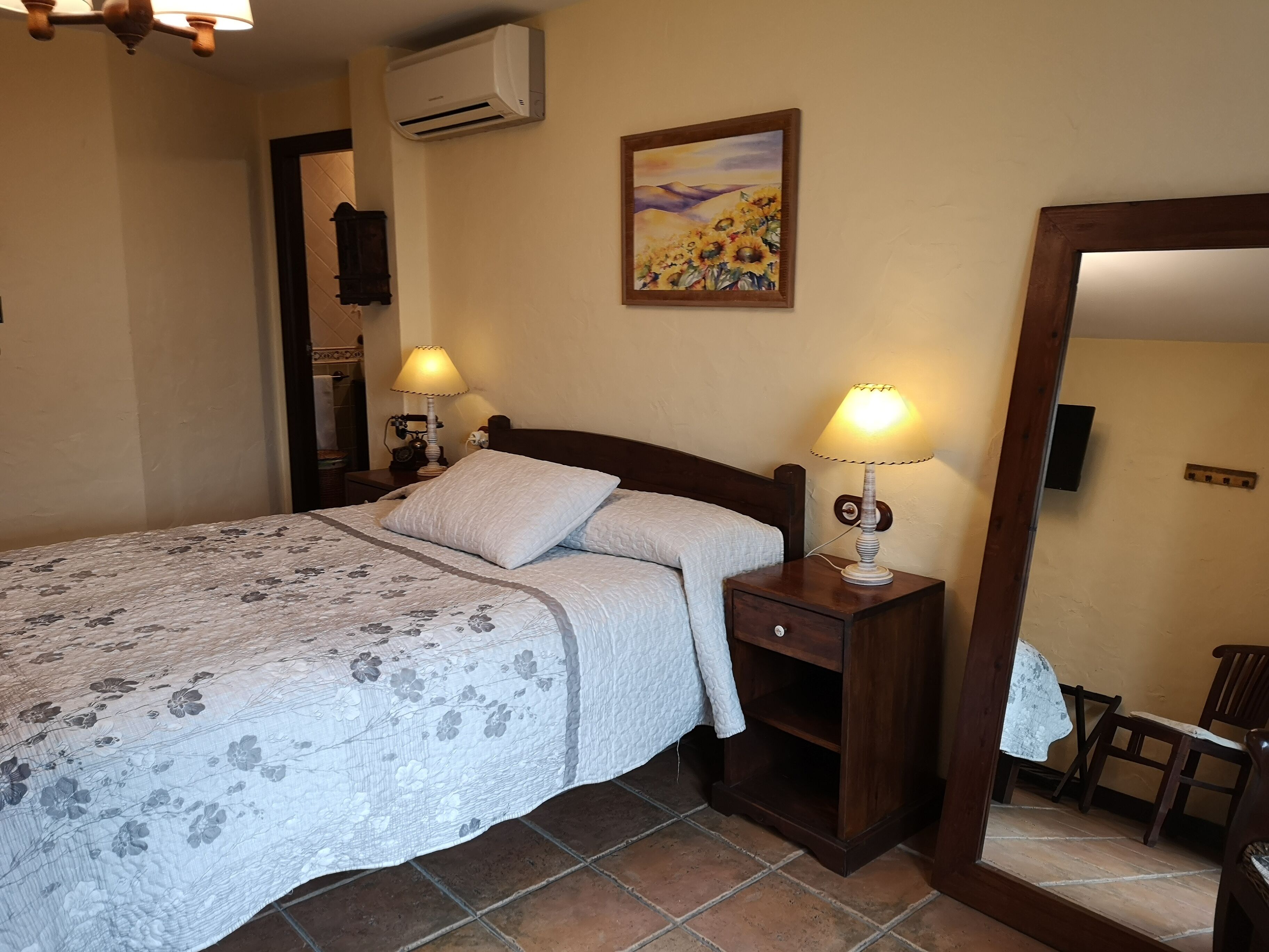 A bedroom with a bed, two bedside lamps, a wall-mounted air conditioner, a painting, a mirror, and a chair.