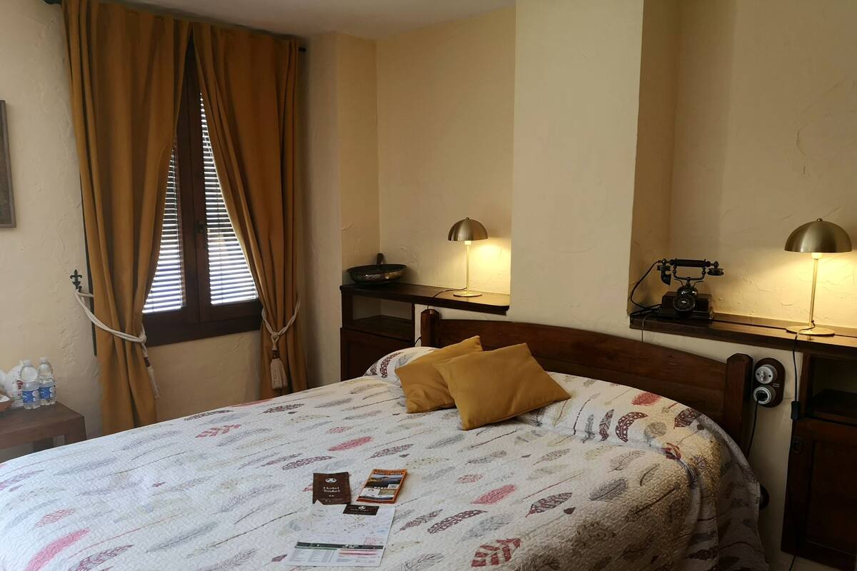Quarto de hotel com cama, duas luminárias de cabeceira, mesa de cabeceira de madeira, espelho e janela com cortinas.