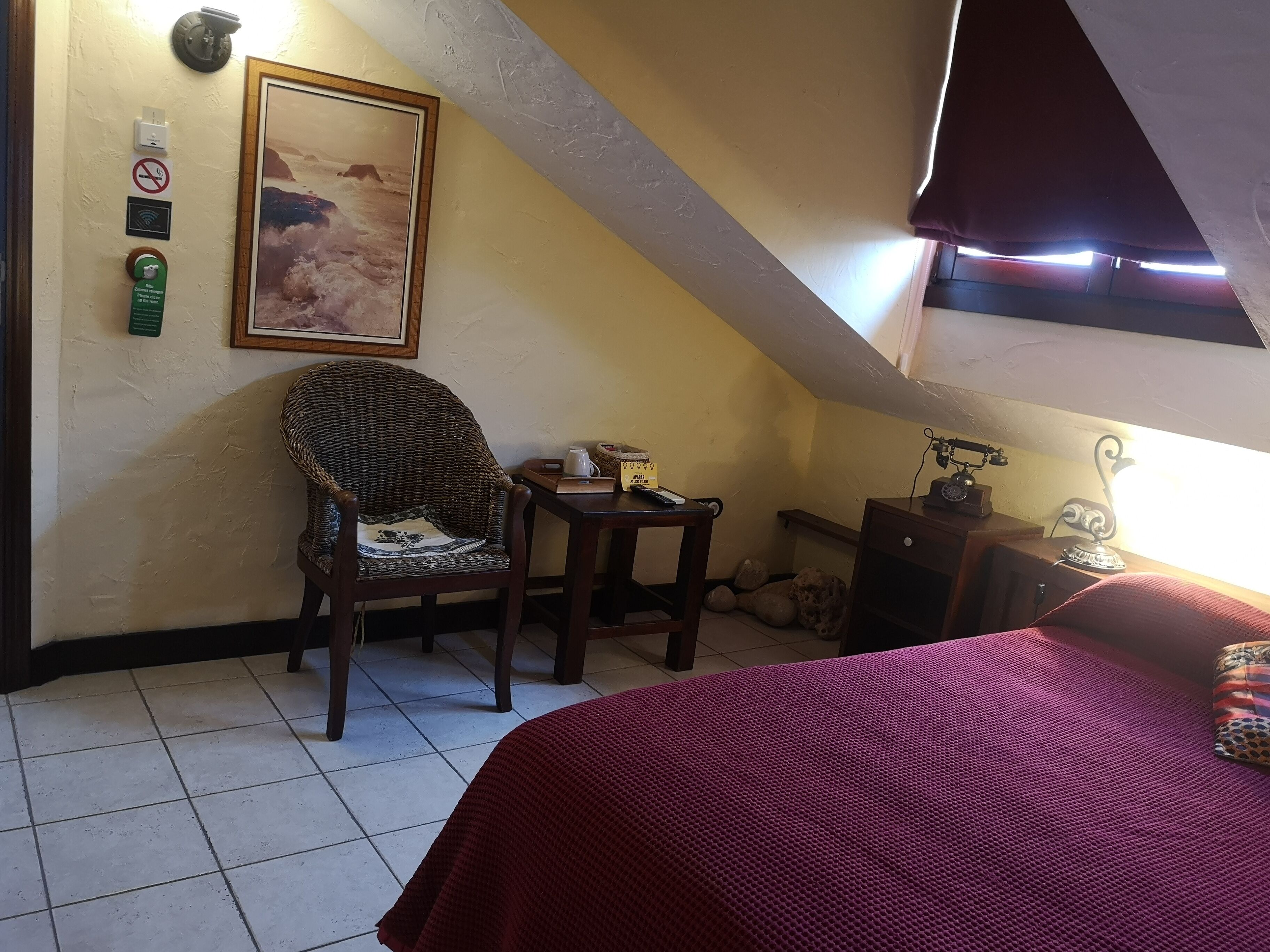 Una stanza accogliente e intima con soffitto spiovente, una poltrona in vimini, un tavolino con un bollitore, un letto con coperta rossa e una finestra con tende.