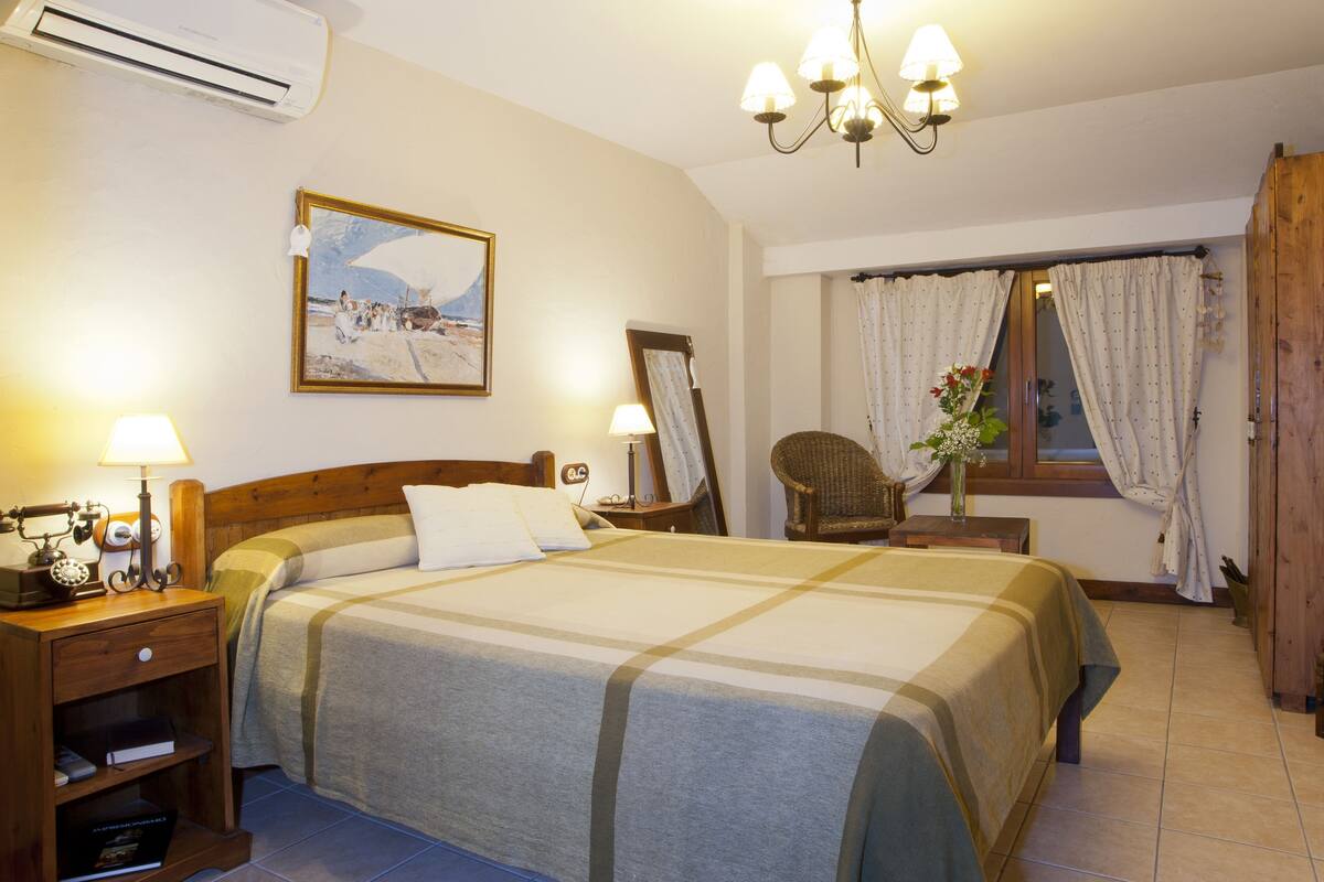 Quarto de hotel com cama, mesa de cabeceira com luminária, cadeira, janela com cortinas e um quadro na parede.