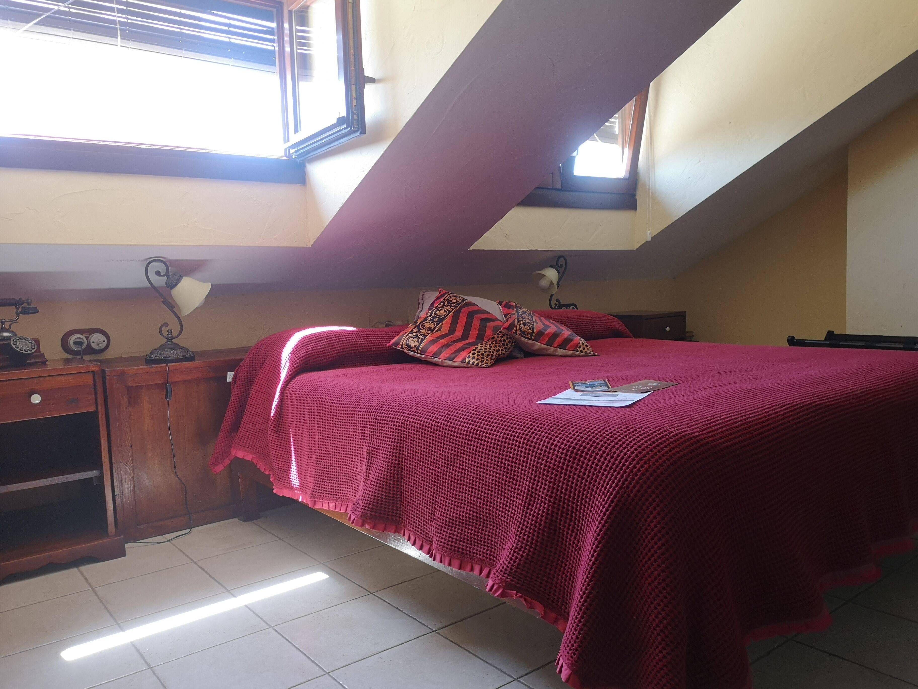 A bedroom with a sloped ceiling, a bed with a red blanket, a desk with a lamp, and a window with blinds.