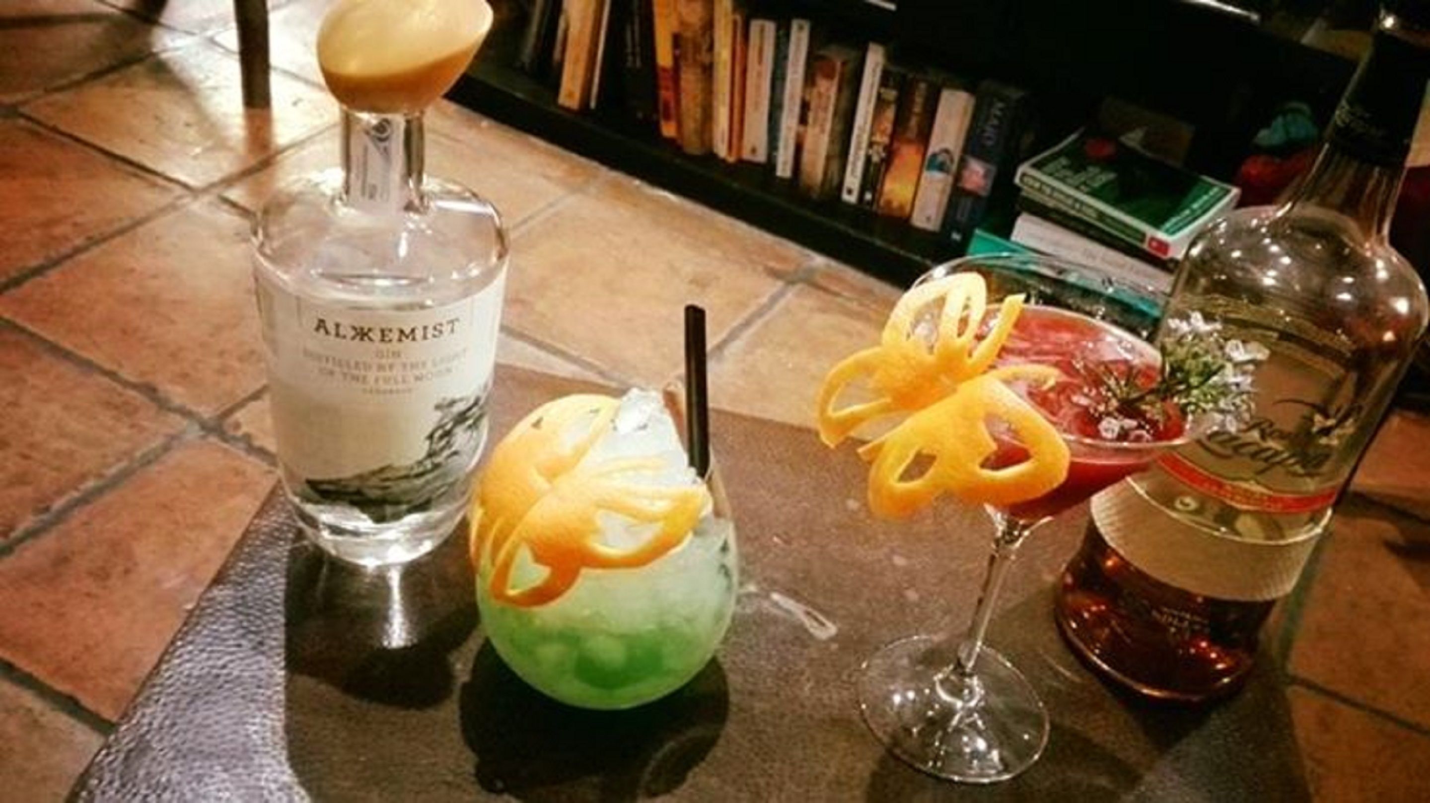 A bar area with a bottle of Alkemist gin, a cocktail with a lime garnish, and a bottle of Campari.