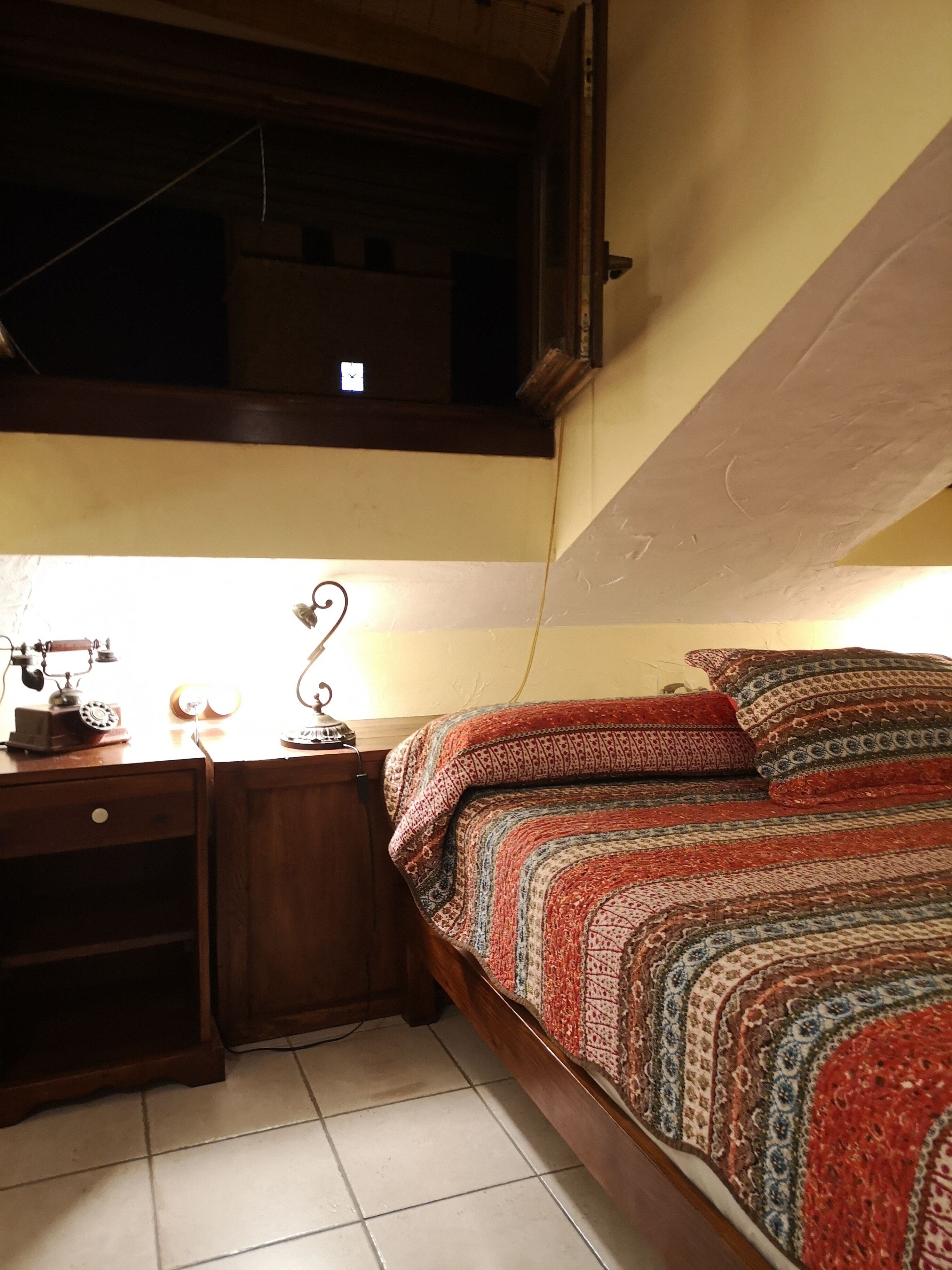 A bedroom with a bed, a desk with a lamp, and a window.