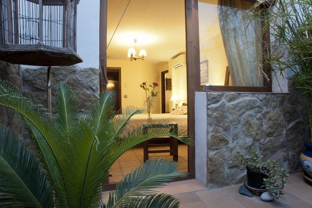 Uma parede de pedra, uma cadeira de madeira, uma planta em vaso e uma cama com uma mesinha de cabeceira.