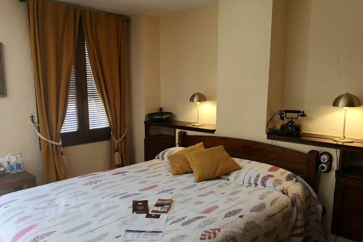 Quarto de hotel com uma cama, duas luminárias laterais, uma mesa de cabeceira de madeira com uma luminária, um espelho e uma janela com cortinas.