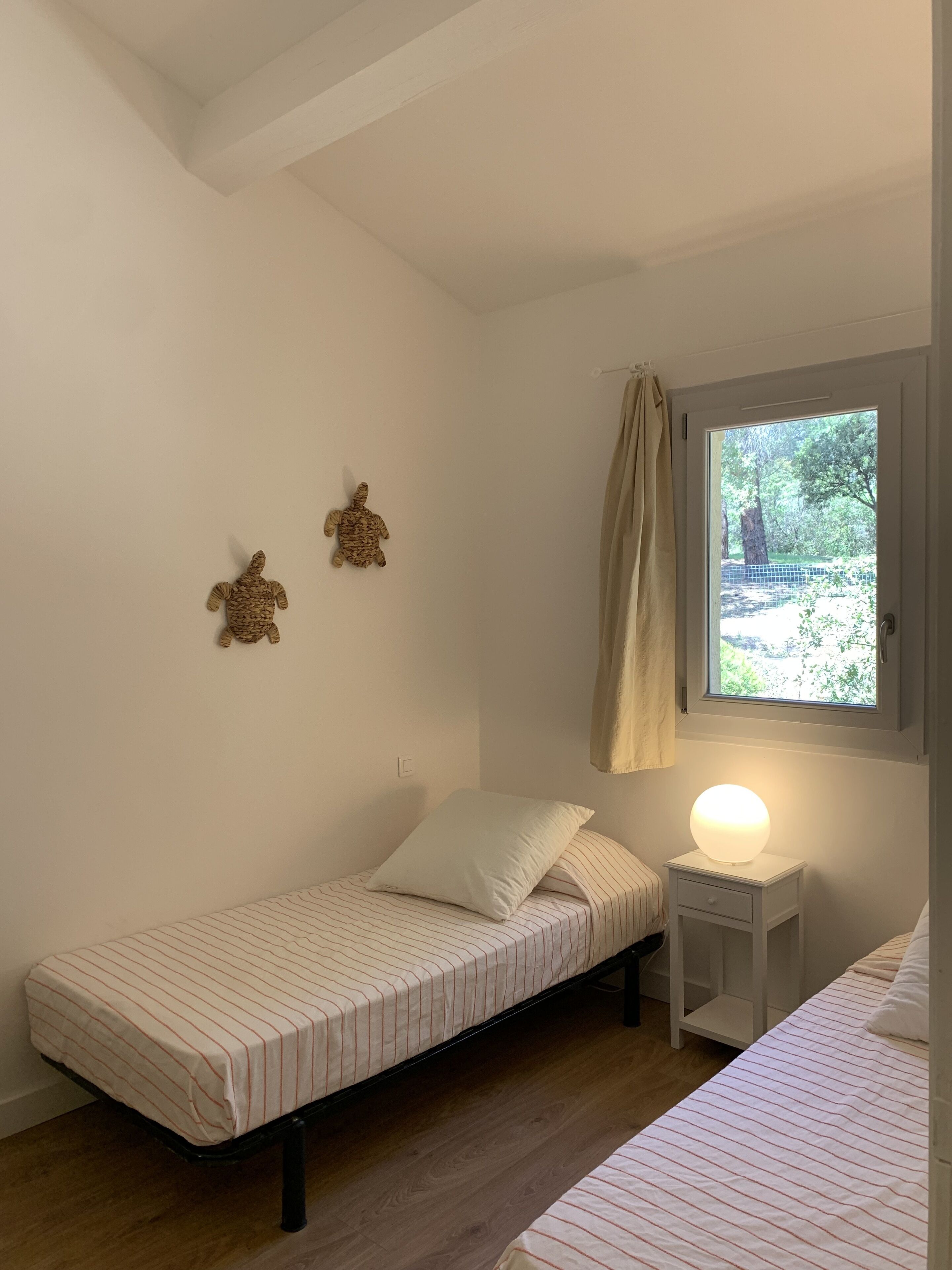 A small, single-bed room with a window, a bedside table with a lamp, and two wall decorations.
