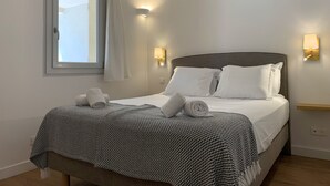 A neatly made bed with white linens and a patterned throw, a window with a curtain, and wall-mounted light fixtures. - Les Pins de Santa Giulia (Porto-Vecchio)