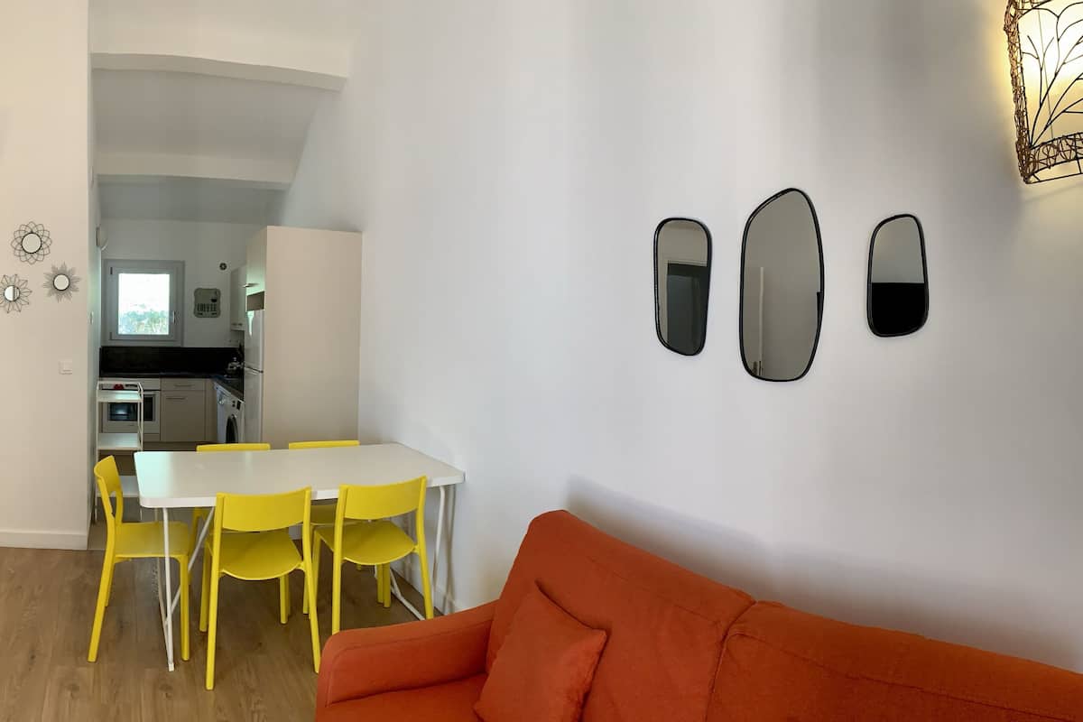 Uma sala de estar moderna com televisão, sofá, espaço de refeições com cadeiras amarelas e cozinha ao fundo.