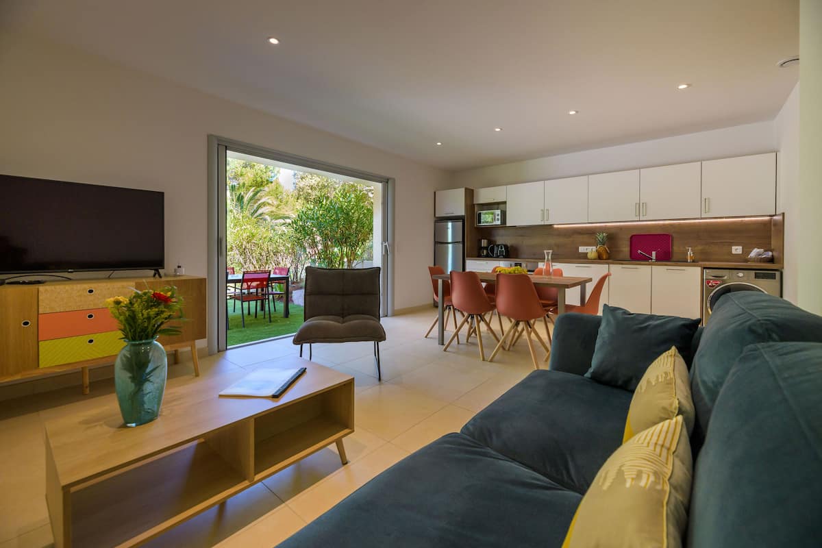 Uma sala de estar moderna com um sofá, uma mesa de centro e uma televisão. Há uma área de jantar com cadeiras e uma mesa, e uma cozinha com armários e eletrodomésticos.