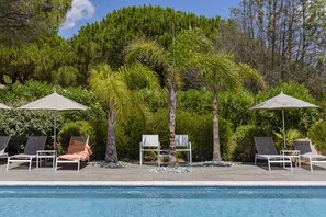 A poolside area with lounge chairs and umbrellas, surrounded by lush greenery and trees.