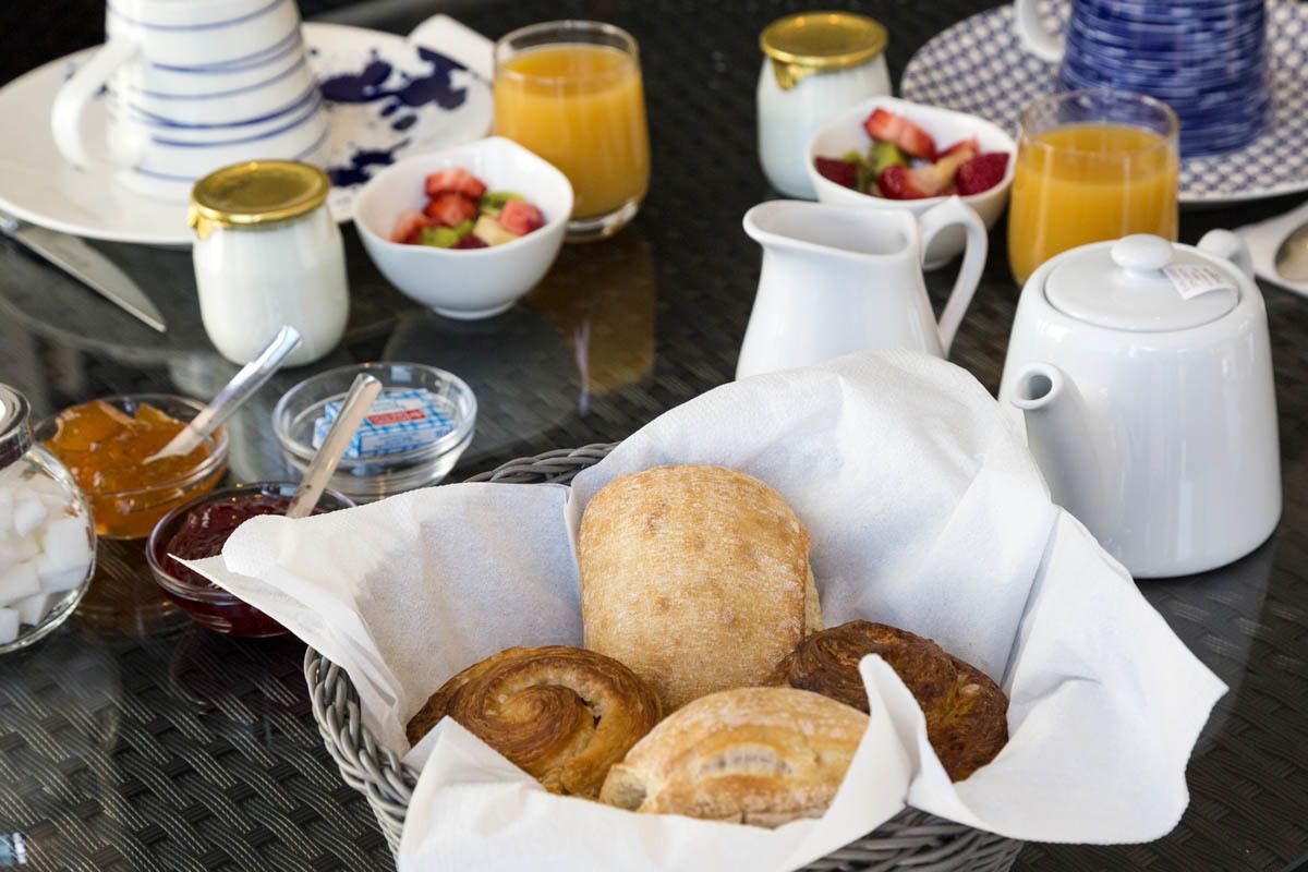 Desayuno con pastelería, una cafetera y cuencos de fruta sobre una mesa.