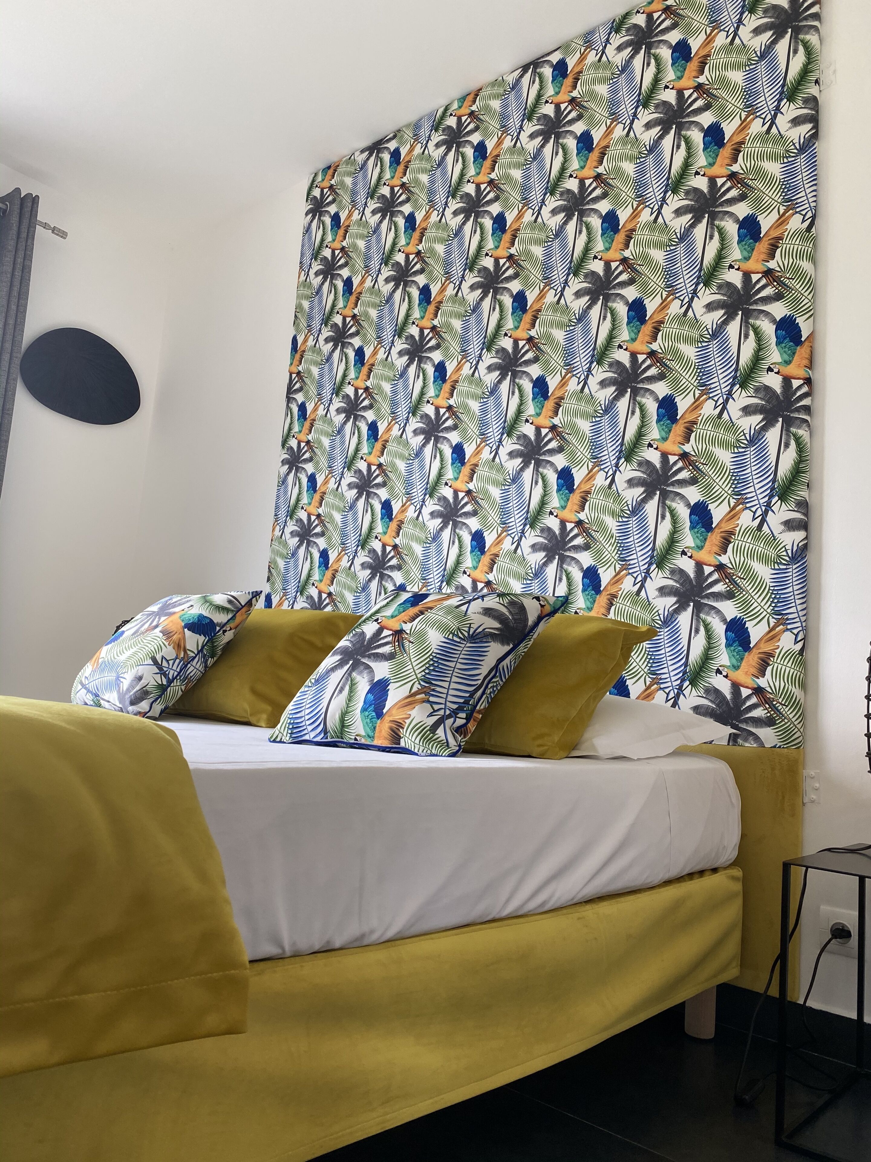 Un lit avec une tête de lit jaune et des oreillers, une couvre-lit blanche et un mur orné d’un motif représentant un oiseau tropical et des feuilles de palmier.