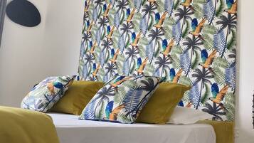 Un lit avec une tête de lit jaune et des oreillers, une couvre-lit blanche et un mur orné d’un motif représentant un oiseau tropical et des feuilles de palmier.