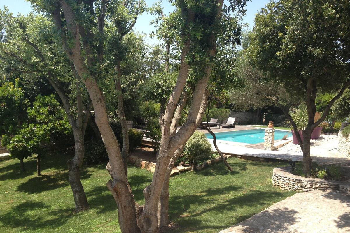 Un jardin à l’arrière de la maison, avec une piscine, entouré d’arbres et bordé d’un mur en pierre.