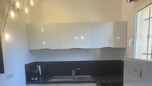 Uma cozinha moderna com armários brancos, bancada preta e pia de aço inoxidável. Há uma geladeira à direita e uma cafeteira à esquerda.